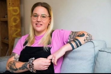 Kaitlin Mountain, wearing a pink shirt, black overalls and tattoos on her arms, leans back on a couch smiling at the camera.