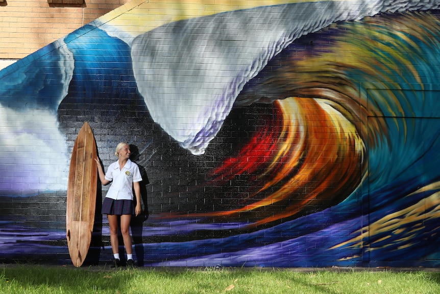 Amira holds a surf board up and looks up at a large crashing wave mural, she is wearing her school uniform.