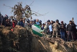 People standing on the edge of a cliff looking downwards. There is plane debris in the foreground. 