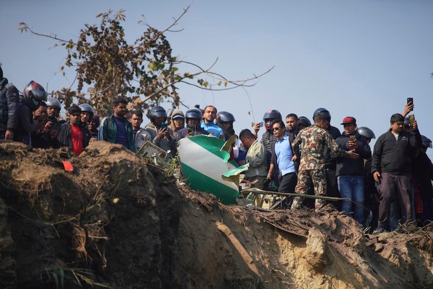 People standing on the edge of a cliff looking downwards. There is plane debris in the foreground. 