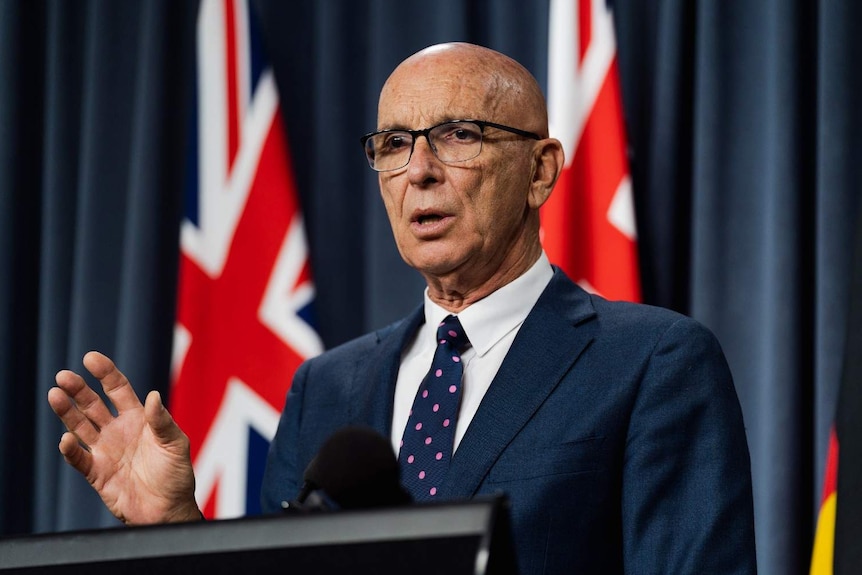 A close up of Attorney-General John Quigley wearing a blue suit and tie standing in front of Australian flags and a blue curtain