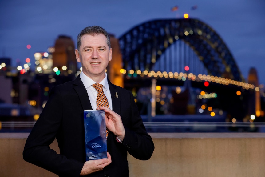 A man in a suit holds a glass award in front of the Sydney Harbor Bridge.