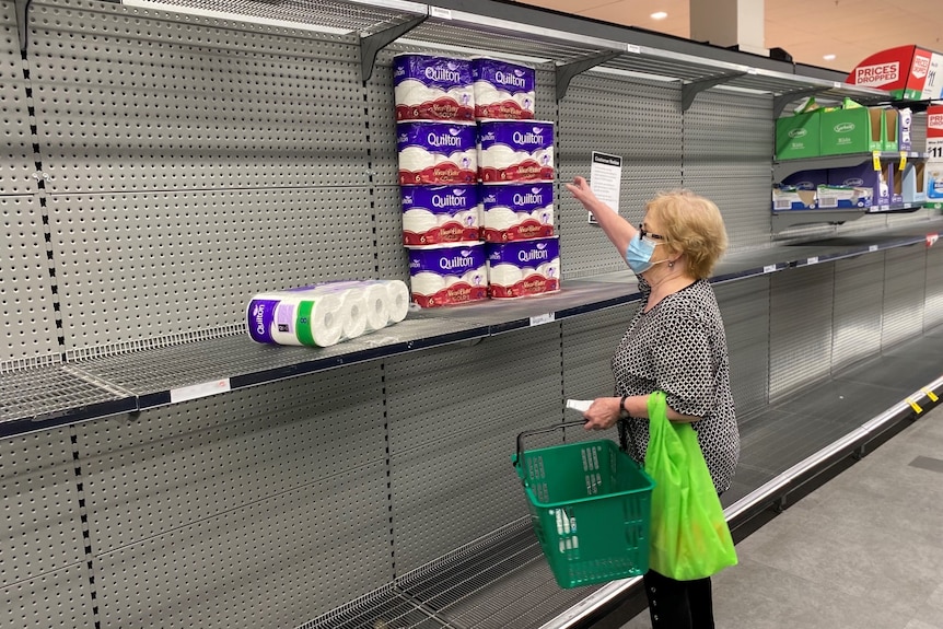 Woman reaching for a stack of toilet paper sitting in the middle of an otherwise empty supermarket shelf.