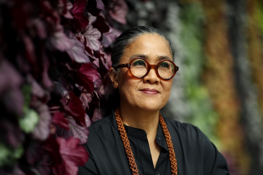 Kylie Kwong wears red thick-rimmed glasses and a black top, looking out to distance standing against a wall of plants + foliage