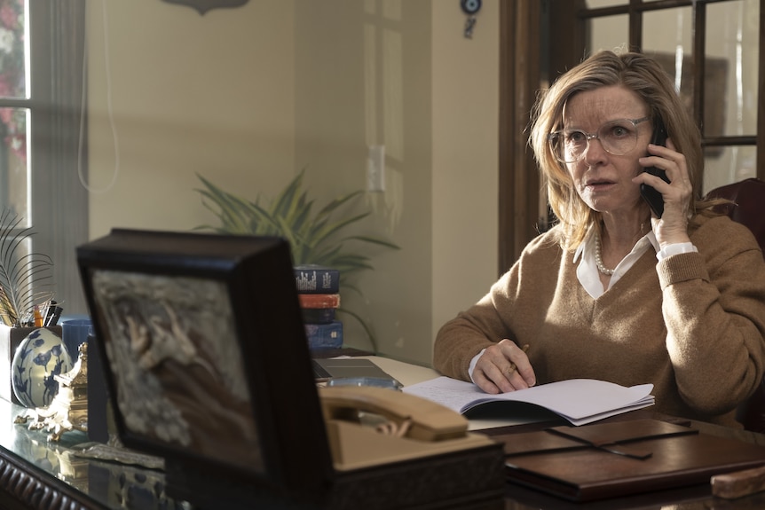 A woman in her 50s with big glasses sits a desk, she has a phone up to her ear and looks concerned