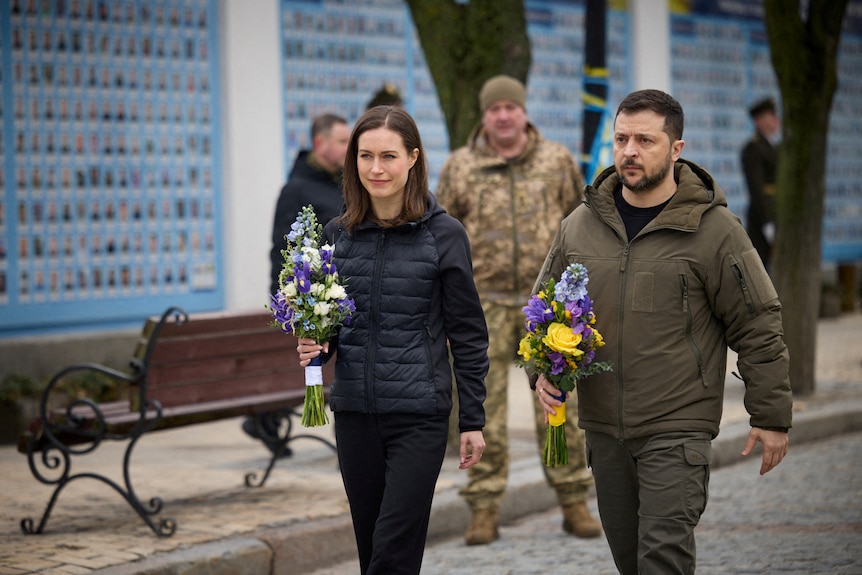 Marin walks beside Zelenskyy, both holding flower bouquets. A uniformed soldier stands in the background