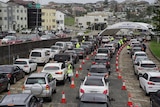 Cars queuing at a drive-through testing clinic on an overcast day with buildings in the background