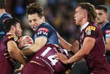 A NSW male State of Origin player holds the ball as he is tackled by three Qld players.