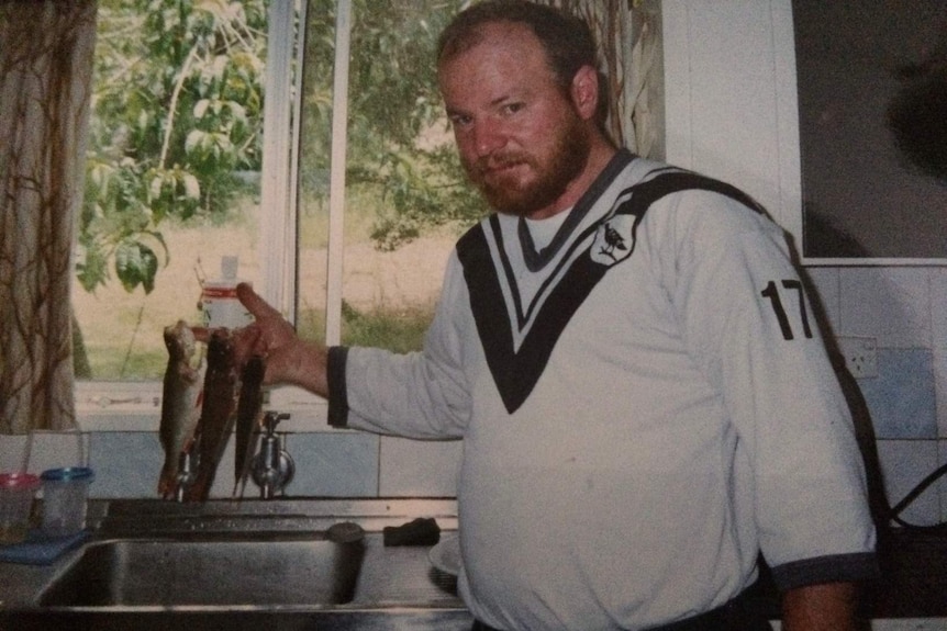 David Wilson stands in a black and white rugby jersey dangling three fish over a kitchen sink