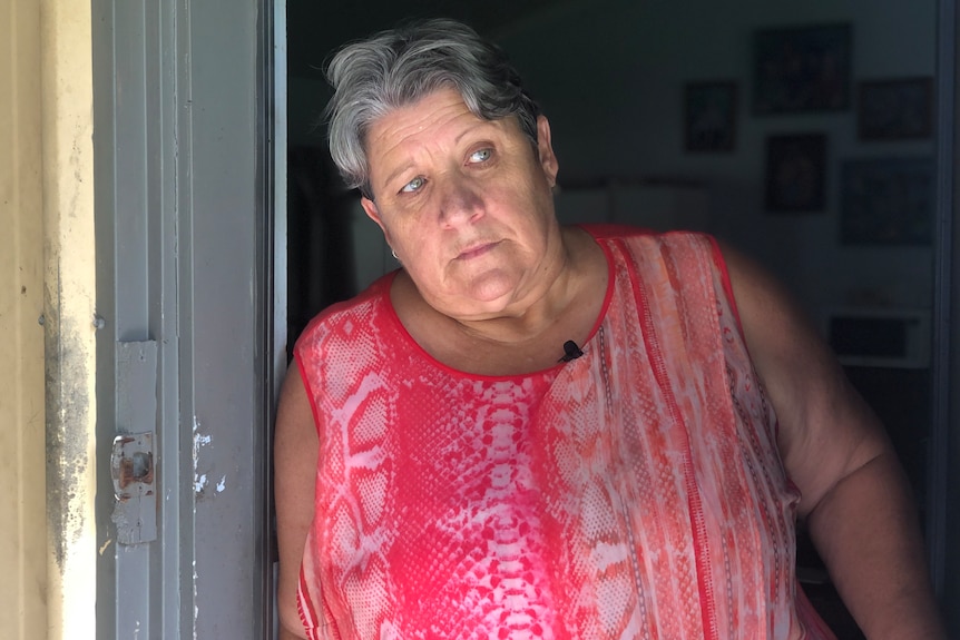 An older woman stands at her door, peering outside, looking worried