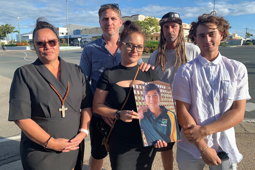 Two women and three men standing side by side in front of a roundabout, holding a framed photo.