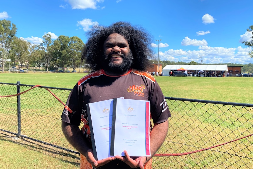 A smiling Indigenous man with a beard and long hair stands in front of a sports field, holding native title documents.