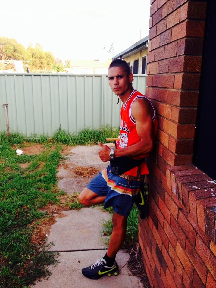 A young man stands near a brick wall giving the thumbs up signal