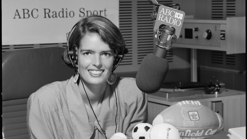 Black and white photo of a woman behind a microphone surrounded by various balls and sporting equipment.