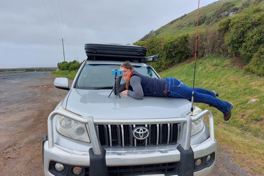 A woman on the bonnet of a four-wheel drive, setting up a small phone tripod.