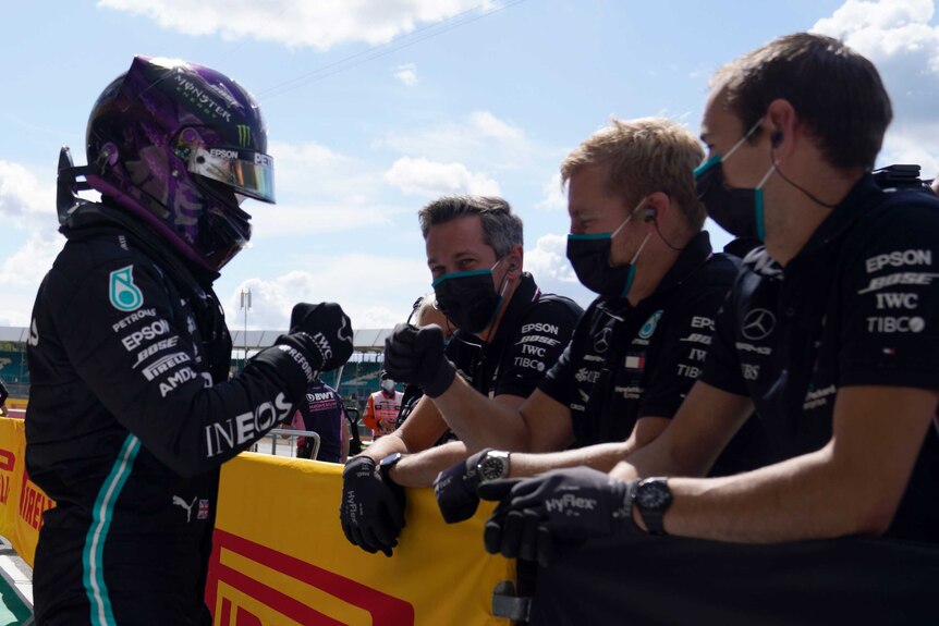 An F1 driver with helmet on bumps fists with his mask-wearing crew at qualifying for a grand prix.