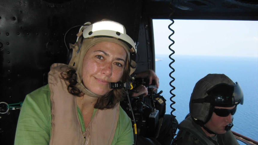 Lisa Millar looks calm while sitting next to military in a helicopter which has its door open.