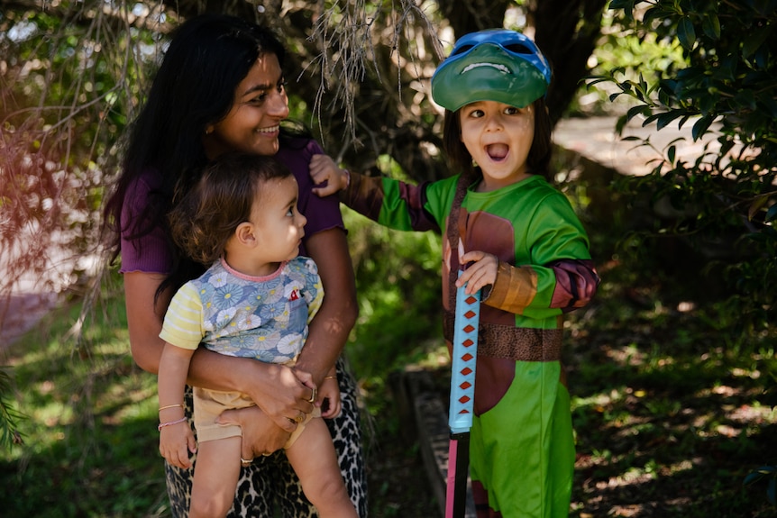 A woman holding a small child, smiling at another child dressed in a Ninja Turtles outfit.