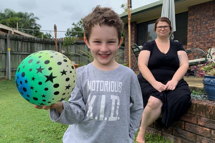 A boy smiles while holding a colourful bouncy ball in the backyard. His mother sits in the background, looking on.