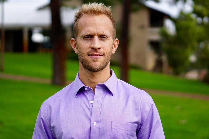 David Lewis stands in a green space. He wears a purple collared shirt. Background of tree trunks and a building and grass.