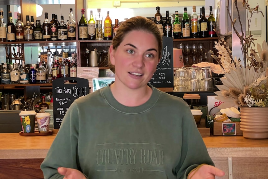 Angela Nichols in a green sweater in from of the bar at Raincheck Lounge. Bottles of alcohol sit on shelves behind her.