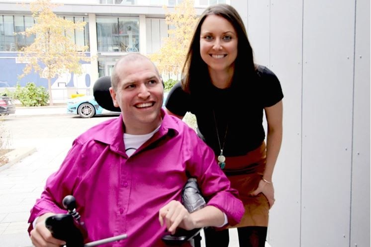 A young man in a wheelchair wearing a bright pink shirt stands next to a young woman in a black tshirt