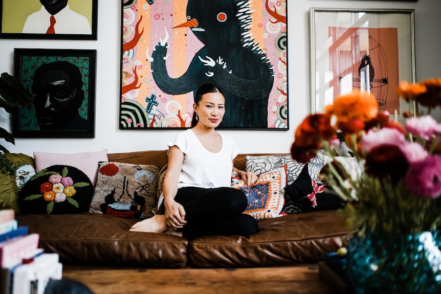 A woman sits with her feet underneath her on a leather couch surrounded by bright artwork.