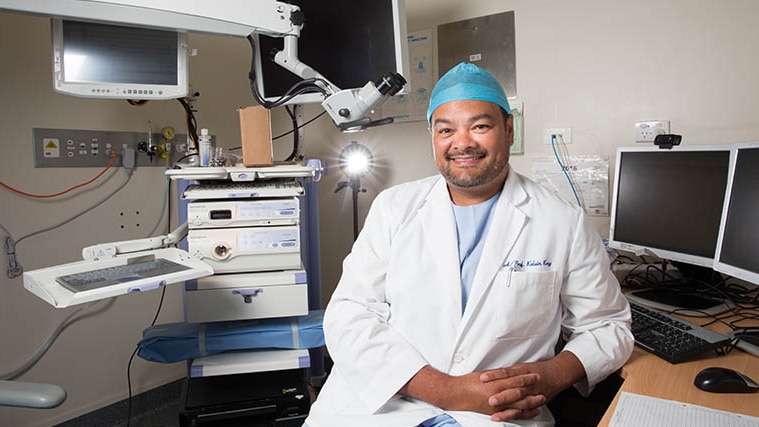 An Indigenous man wears hospital scrubs, cap, white jacket, surrounded by medical equipment and smiles at camera.