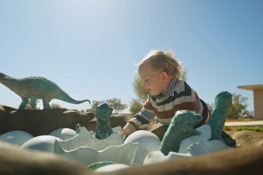 A blonde toddler sits in a dinosaur nest sculpture. 
