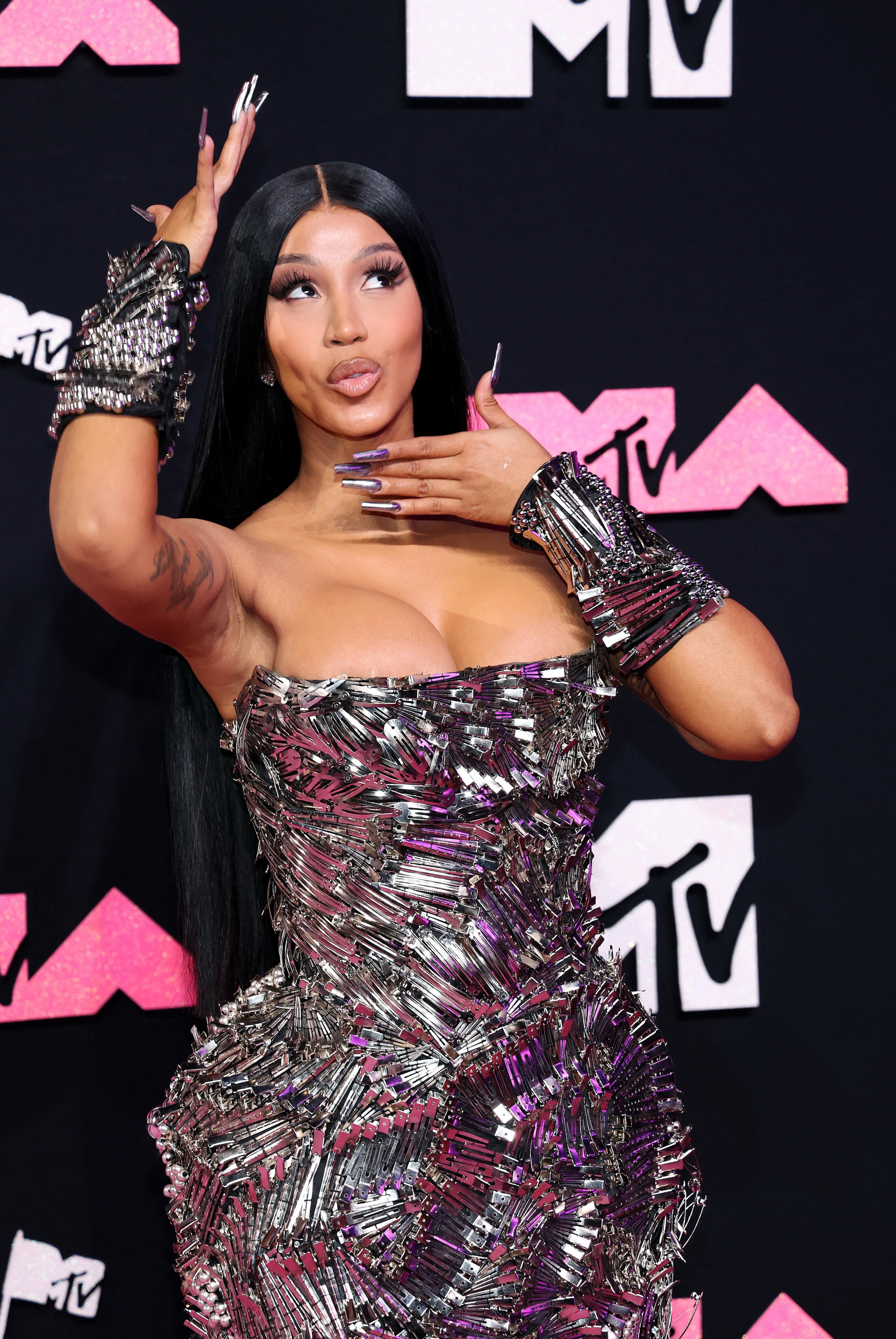 Cardi B is wearing a shiny silver dress while she poses holding her hands up to her face.