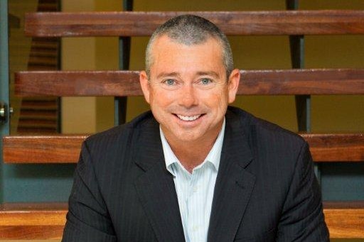 A middle-aged man in a black suit sitting on bleaches smiles at the camera.