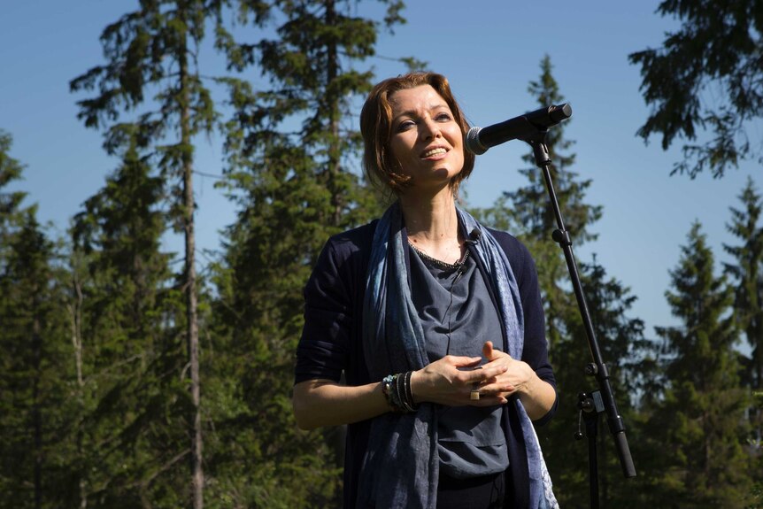 Brown-haired woman in grey top with blue scarf draped around her neck stands in front of a microphone in the middle of a forest.