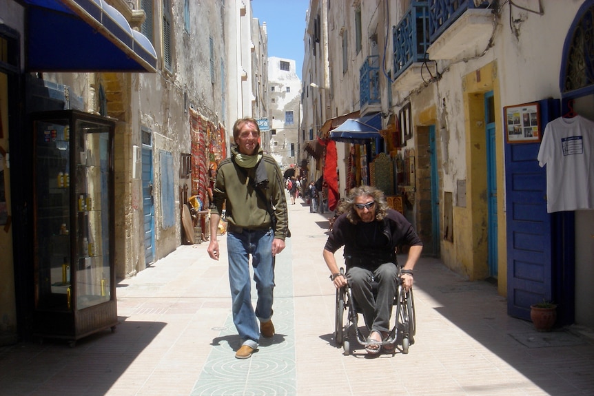 Man walking next to man in a wheelchair in a Morrocan street lined with coloured doors and shop fronts, no people.
