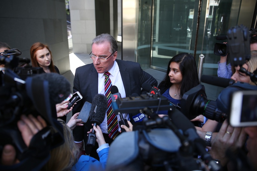 A man in a suit and a young woman are speak to a media pack on a street.