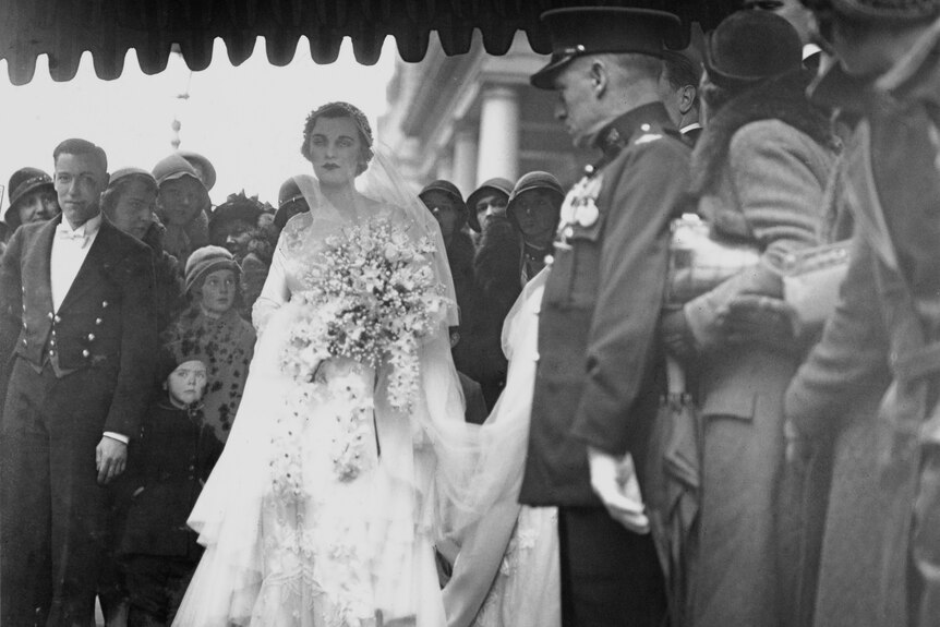 A woman wearing a white wedding dress and holding a bouquet of flowers stands surruonded by men in suits.