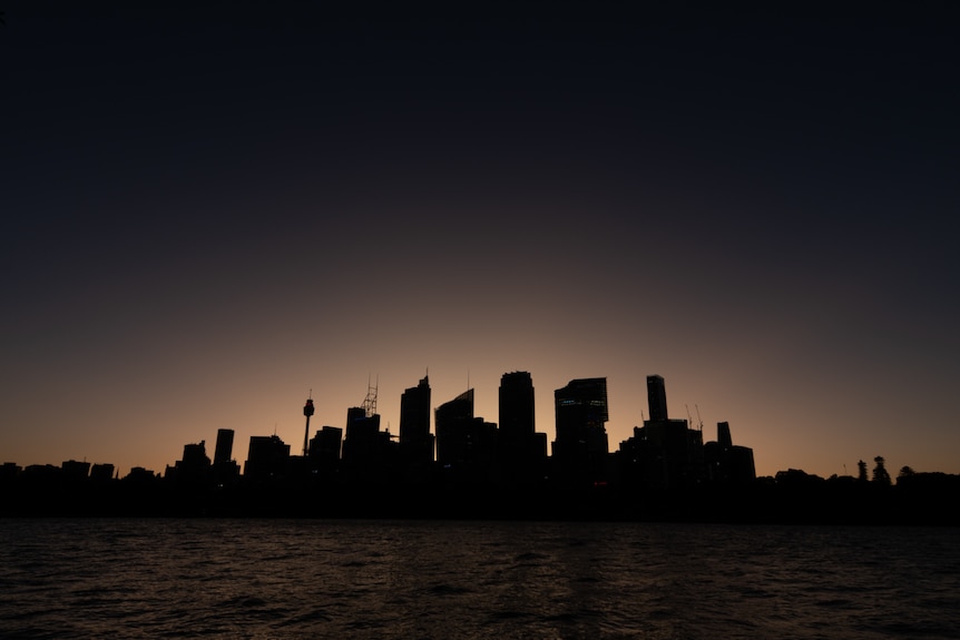 Sydney skyline in darkness, orange glow behind, taken across water.