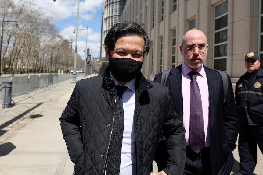 Roger Ng in a jacket and tie and black face mask walks next to a man in a suit along a street outside a courthouse