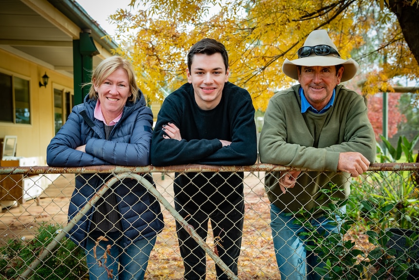 A young man stands between his parents, leaning up against an old fence. A tree with yellow leaves fills the background