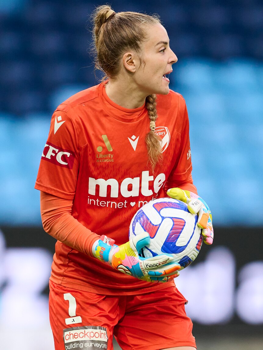 A soccer goalkeeper wearing orange holds a ball in her colourful gloves during a game