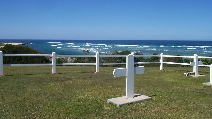 Two white crosses on a patch of land overlooking the ocean, surrounded by a white fence.