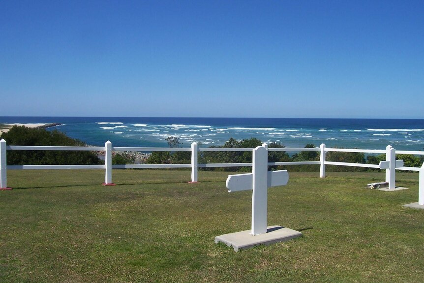 Two white crosses on a patch of land overlooking the ocean, surrounded by a white fence.