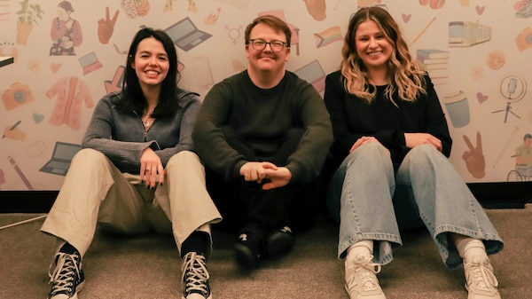 Three young people sit on the floor of an office, leaning against a wall, smiling.