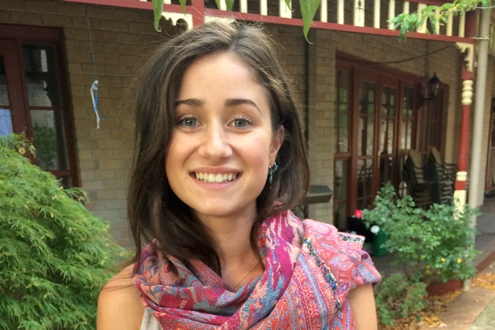 A young woman in a grey singlet smiles and a pink scarf smiles at the camera.