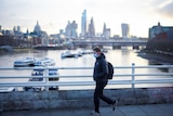 A man in a puffy jacket and face mask walks over a bridge with the London skyline behind him