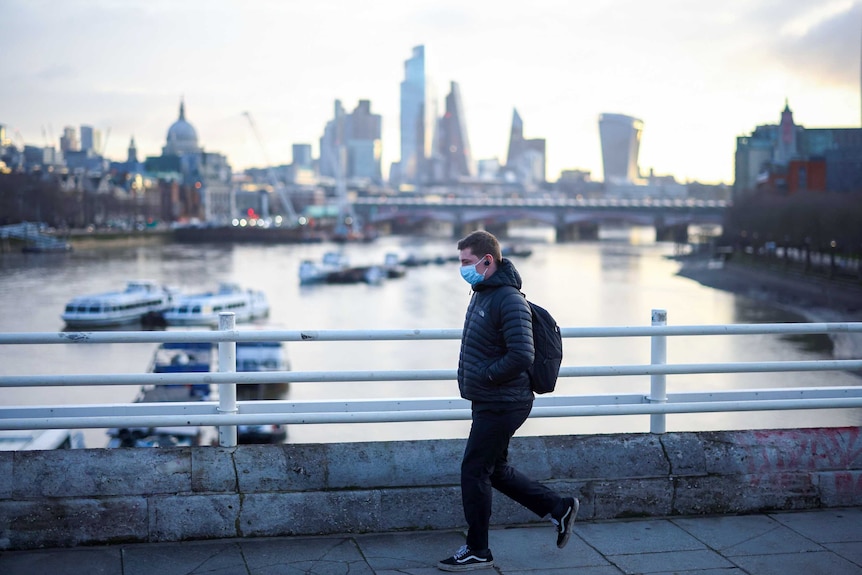 A man in a puffy jacket and face mask walks over a bridge with the London skyline behind him