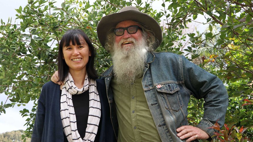 Greg, who wears a felt hat with a fur band, and has a large, rambunctious beard, stands with a smiling Linh in front of a tree.