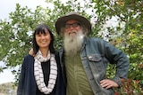 Greg, who wears a felt hat with a fur band, and has a large, rambunctious beard, stands with a smiling Linh in front of a tree.