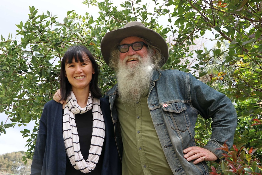 Greg, who wears a felt hat with a fur band, and has a large, rambunctious beard, stands with a smiling Linh in front of a tree.