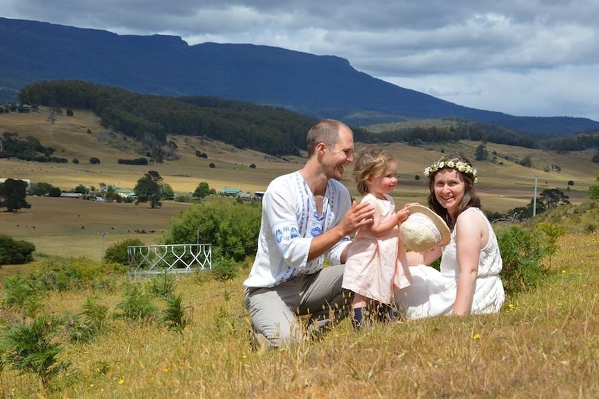 Jenny and Tom with their daughter sit on a hill after their wedding.