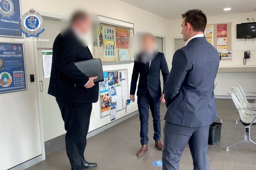 Three men, two with their faces blurred, stand in a police station.